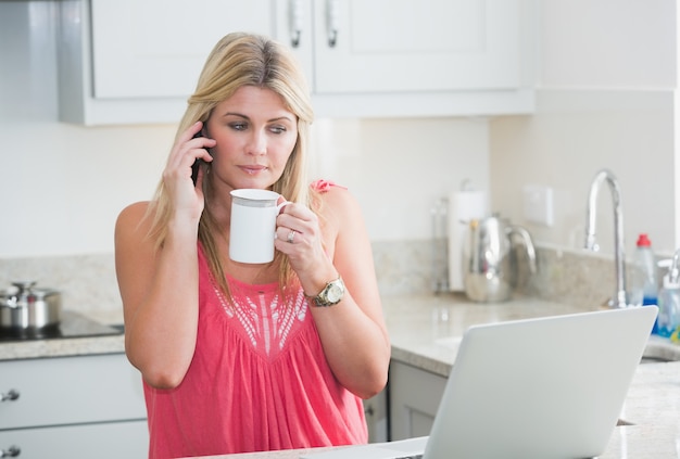 Frau mit Laptop und Kaffeetasse beim Anruf in der Küche