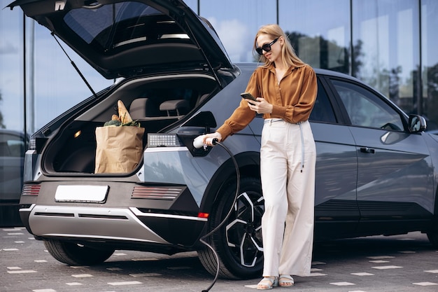 Foto frau mit lebensmittel-shopping-taschen, die ein elektroauto laden und ein handy benutzen