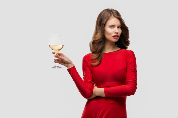 Frau mit Make-up, Frisur, die rotes Kleid trägt, das mit Glas Weinstock über weißer Wand aufwirft, isolieren.