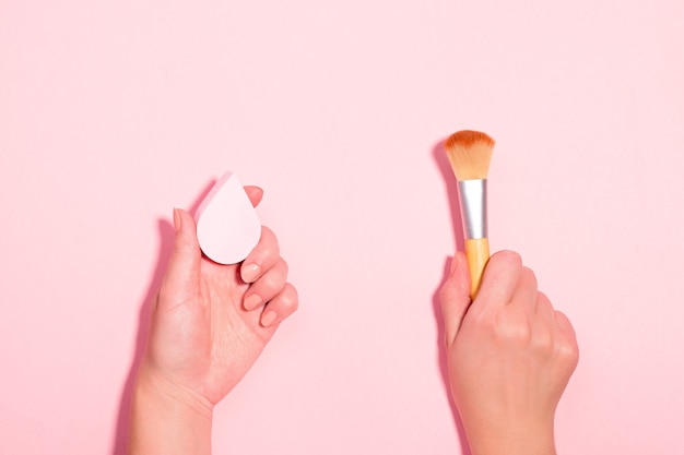 Frau mit Make-up-Pinsel und Beauty-Mixer auf pastellrosa Hintergrund. Beauty-Accessoires mit Make-up-Pinsel versus Beauty-Schwamm für Foundation. Schönheit flach, Ansicht von oben, Kopienraum