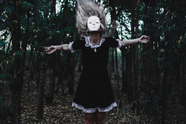Frau mit Maske und ausgestreckten Armen steht im Wald