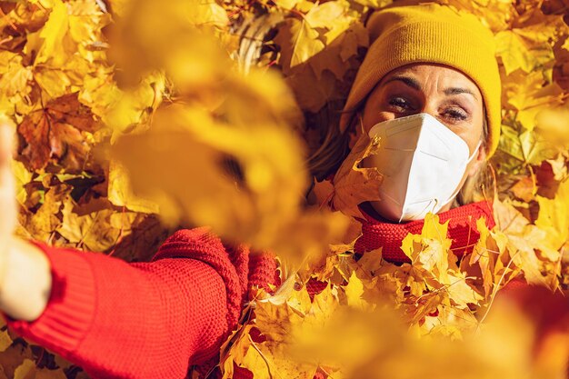 Frau mit medizinischem Virenschutz knN95 FFP2-Gesichtsmaske zur Vorbeugung gegen Coronavirus COVID19 wirft orangegoldene Herbstblätter auf, die von oben auf gelben und orangefarbenen Blättern im Park liegen