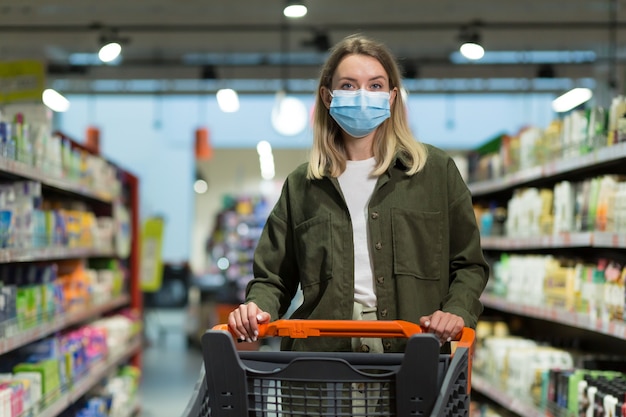 Frau mit medizinischer Gesichtsmaske schiebt Einkaufswagen im Supermarkt. Junges Mädchen, das wählt, Lebensmittelgeschäfte sucht, um zu kaufen. Mädchen geht durch Supermarkt oder Laden. fröhliche Hübsche Frau, die im Einkaufszentrum spazieren geht