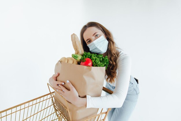 Frau mit medizinischer Maske, die eine Einkaufstasche voller frischer Lebensmittel hält.