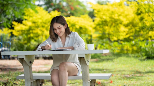 Frau mit nachdenklichem Lächeln im Park mit Notizbuch und Schreiben Porträt einer jungen charmanten Geschäftsfrau, die online überprüft Geschäftliche Arbeit an ihrem Smartphone im Freien im Park auf weichem grünen Hintergrund