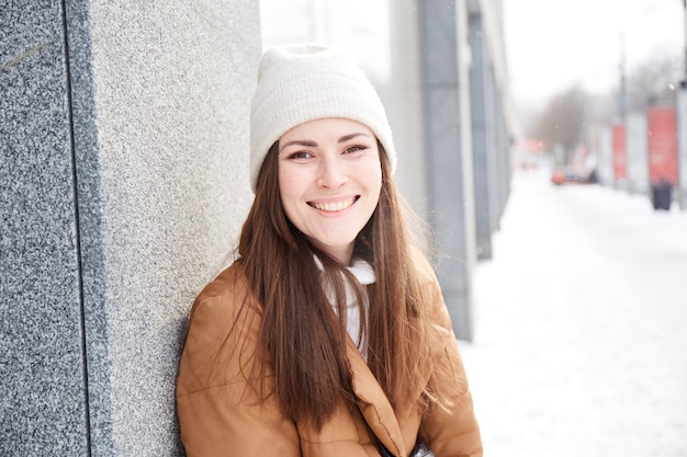 Frau mit natürlichen Sommersprossen, die den Winter auf der Straße in der Stadt genießen. Winterporträt mit Schneeflocken. Tausendjährige brünette Frau im trendigen Outfit