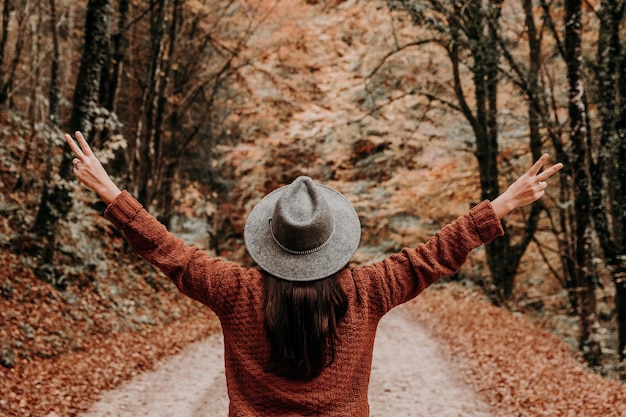 Frau mit offenen Armen, die eine braune Jacke mit Fedora-Hut in einem Wald im Herbst trägt