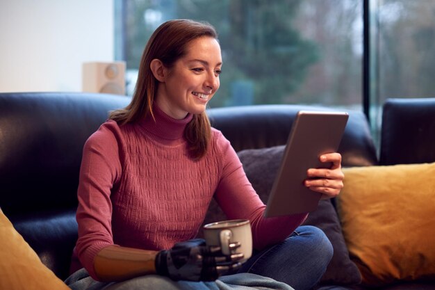 Frau mit prothetischem Arm und Hand zu Hause mit digitalem Tablet-Trinkbecher Kaffee