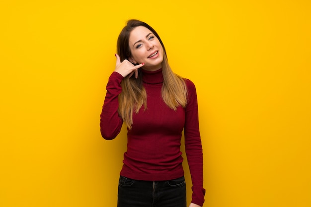 Frau mit Rollkragen über der gelben Wand, die Telefongeste macht. Ruf mich zurück Zeichen