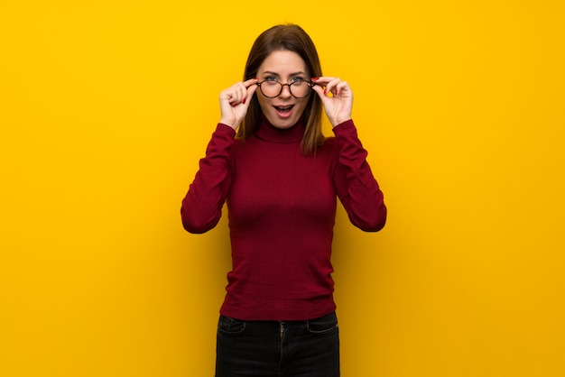 Frau mit Rollkragenpullover über gelbe Wand mit Brille und überrascht