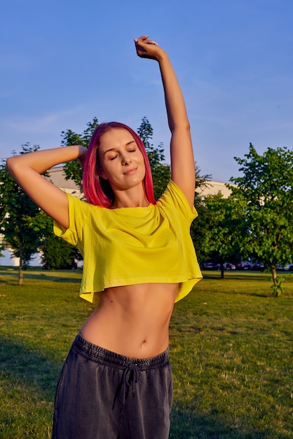 Frau mit rosa Haaren, gelbem bauchfreiem Oberteil dehnt sich vor dem Training im Stadtpark aus
