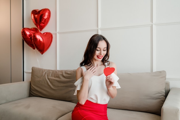 Frau mit rotem Herzen formte die Valentinsgrußkarte, die zu Hause auf Couch mit Ballonen sitzt