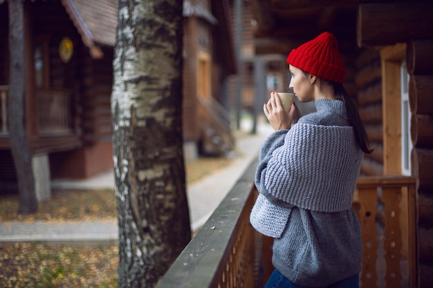 Frau mit rotem Hut und Schal und einem Becher steht an einem Holzhaus im Wald