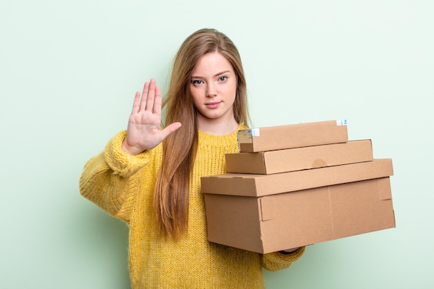 Foto frau mit roten haaren, die ernst aussieht und offene handfläche zeigt, die stoppgeste macht. pakete boxen konzept