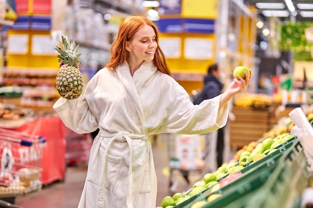 Frau mit roten Haaren, die frisches Gemüse und Obst im Supermarkt kaufen, stehen im Bademantel, genießen das Einkaufen und vergleichen Lebensmittellebensmittelgeschäft