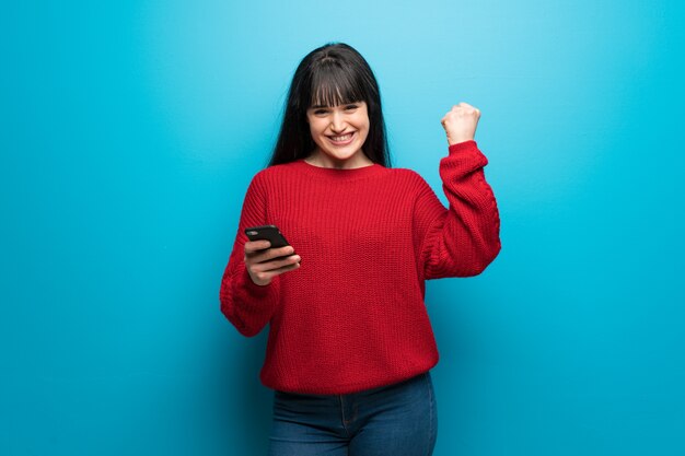 Frau mit roter Strickjacke über blauer Wand mit Telefon in Siegposition