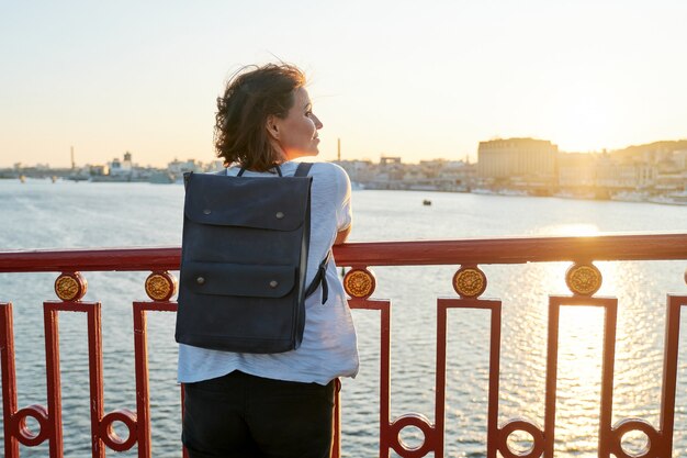 Frau mit Rucksack, die mit Rucksack auf der Brücke steht, Frau, die goldene Stunde genießt, Kopienraum, Sonnenuntergangflusslandschaft, Sommerurlaub, Natur in der Stadt