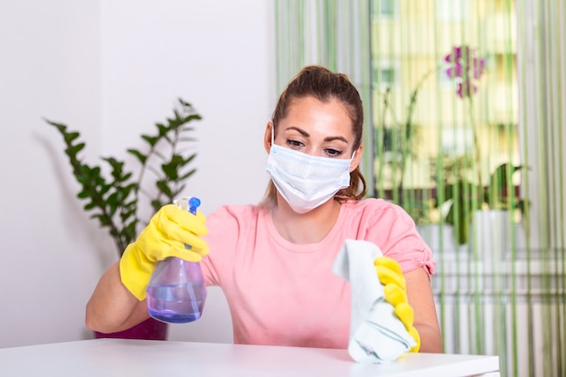 Frau mit Schutzhandschuh und Gesichtsmaske, die Desinfektionsmittel besprüht und den Tisch reinigt. Bleib sicher.