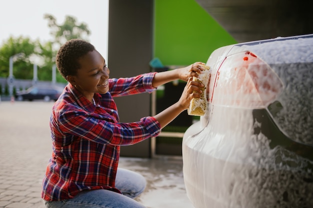 Frau mit Schwamm mit Schaum, Handwaschanlage. Autowaschindustrie oder -geschäft. Weibliche Person reinigt ihr Fahrzeug im Freien von Schmutz