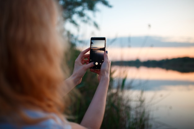 Frau mit Smartphone und Foto des bunten Sonnenuntergangs über dem See.