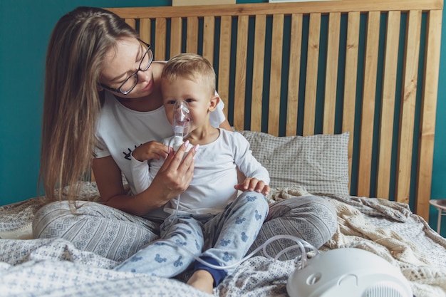 Frau mit Sohn beim Einatmen mit Vernebler in ihrem Schlafzimmer zu Hause