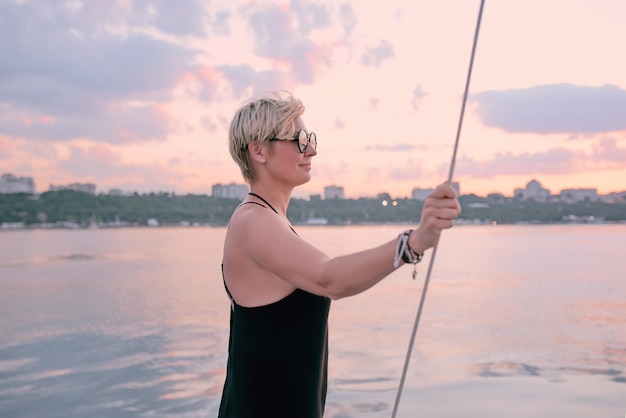 Frau mit Sonnenbrille und langem schwarzem Kleid, die den Sonnenuntergang auf der Yacht im Meer genießt