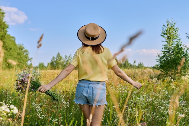 Frau mit Strohhut, die auf der Wiese läuft, mit Strauß wilder Blumen, Rückansicht