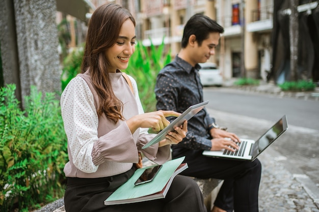 Frau mit Tablet mit jungem Mann, der mit Laptop auf der Parkbank sitzt