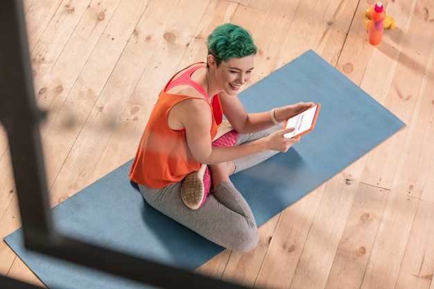 Frau mit Tablette. Grünhaarige Frau in Sportkleidung sitzt auf Sportmatte und benutzt Tablet