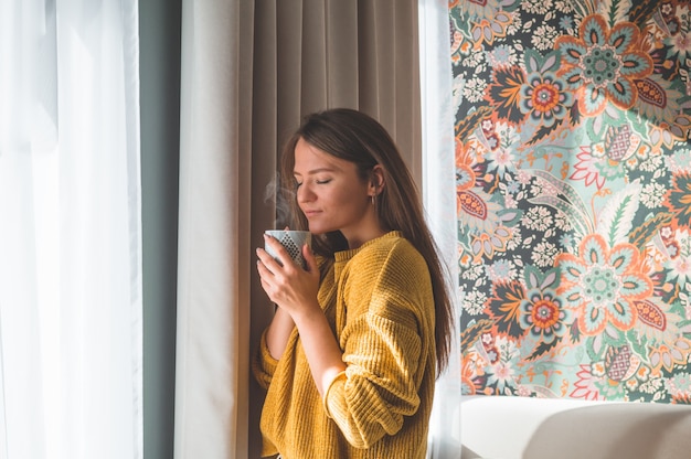 Frau mit Tasse heißem Getränk durch das Fenster. Fenster betrachten und Tee trinken. Guten Morgen mit Tee. Herbst Winter Zeit