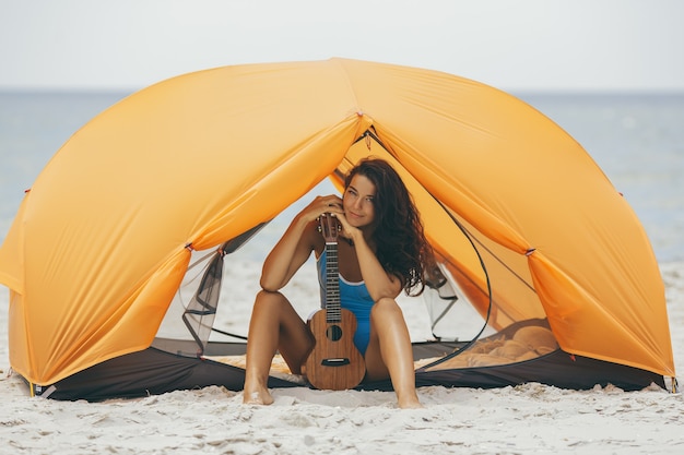 Frau mit Ukulele am Strand unter einem orangefarbenen Zelt