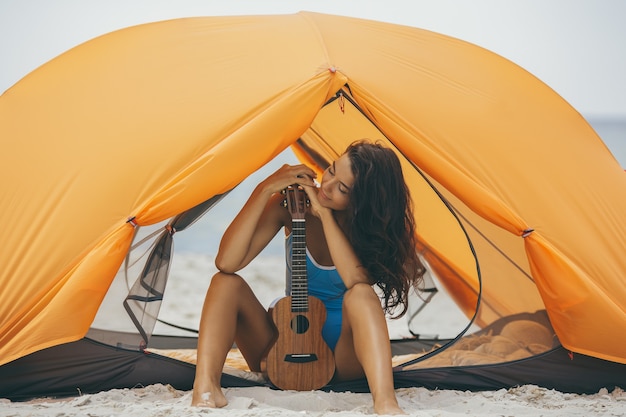 Frau mit Ukulele am Strand unter einem orangefarbenen Zelt