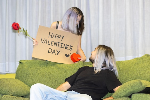 Frau mit Valentines Board geben seiner Freundin auf dem Sofa sitzen ein Geschenk Frau geben seiner Freundin auf dem Sofa sitzen ein Geschenk Valentine