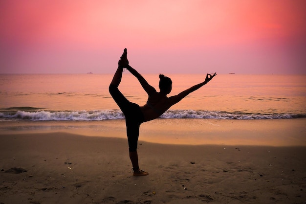 Frau mit voller Silhouette übt Yoga am Strand gegen den dramatischen Himmel während des Sonnenuntergangs
