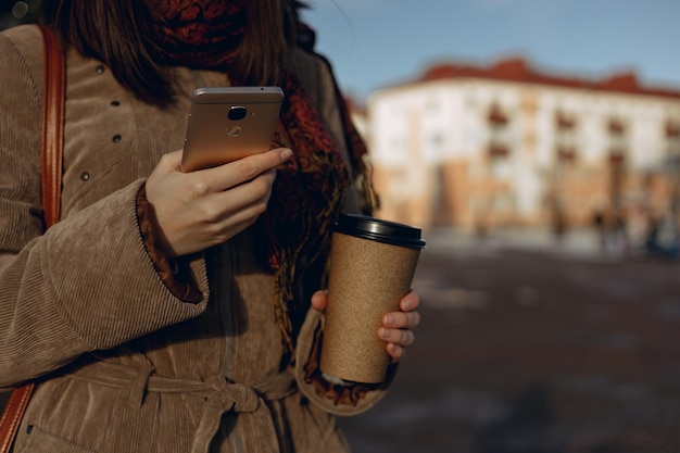 Frau mit warmer Kleidung auf dem Smartphone, während sie mit Kaffee zum Mitnehmen steht