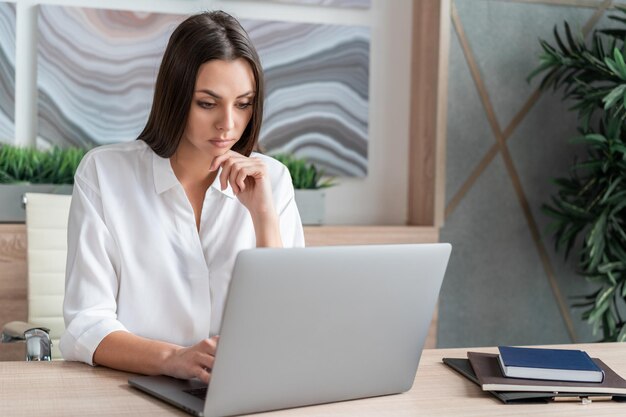 Frau mit weißem Hemd sitzt Tisch mit Laptop und Notizbuch und macht sich Notizen Konzept der Arbeit