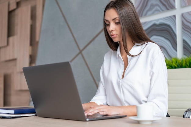 Frau mit weißem Hemd sitzt Tisch mit Laptop und Notizbuch und macht sich Notizen Konzept der Arbeit