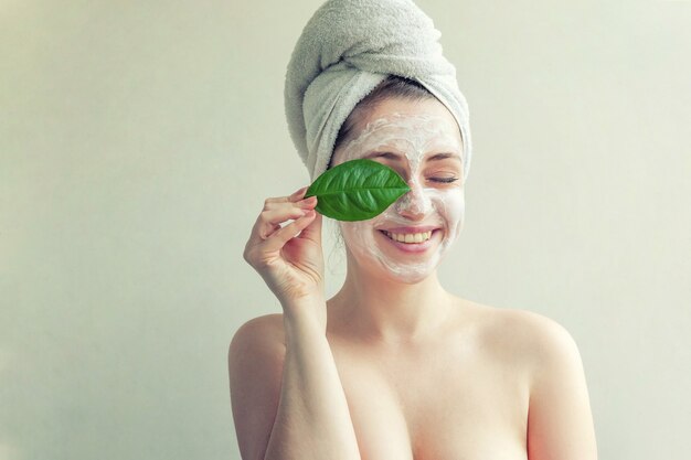 Frau mit weißer pflegender Maske oder Creme im Gesicht und grünem Blatt in der Hand, weißer Hintergrund