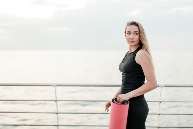 Frau mit yogamatte kommt zum meer, um asana auf sonnenaufgang zu üben