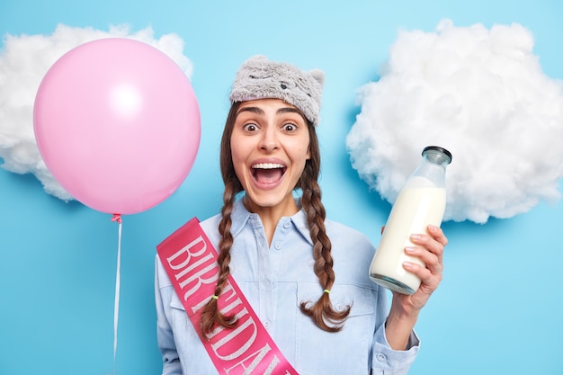 Frau mit Zöpfen hält den Mund offen reagiert auf erstaunliche Neuigkeiten hält eine Flasche frischer Milch aufgeblasener Ballon trägt Schlafmaske auf der Stirn Hemd mit Band geschrieben Geburtstag auf