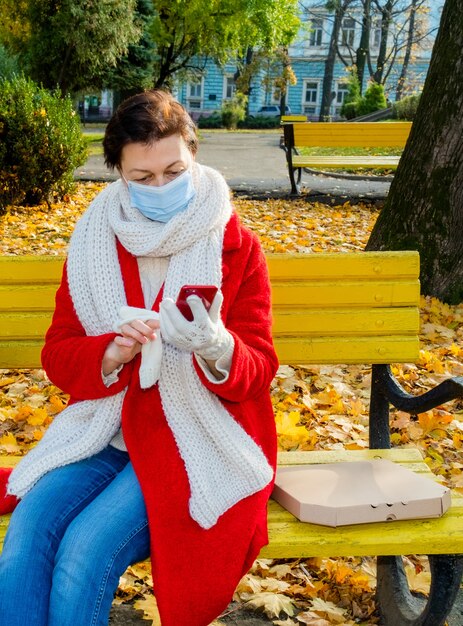 Frau mittleren alters 50 plus mit schützender medizinischer maske, die auf einer gelben bank im herbstpark mit telefon und box für pizza sitzt. liefer- oder mitnahmekonzept.