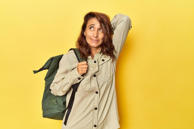 Frau mittleren Alters, bereit für eine Reise mit Rucksack auf gelb, berührt den Rücken des Kopfes und denkt