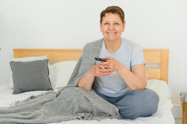 Frau mittleren Alters, die auf dem Sofa sitzt und den Fernseher mit einer Fernbedienung einschaltet