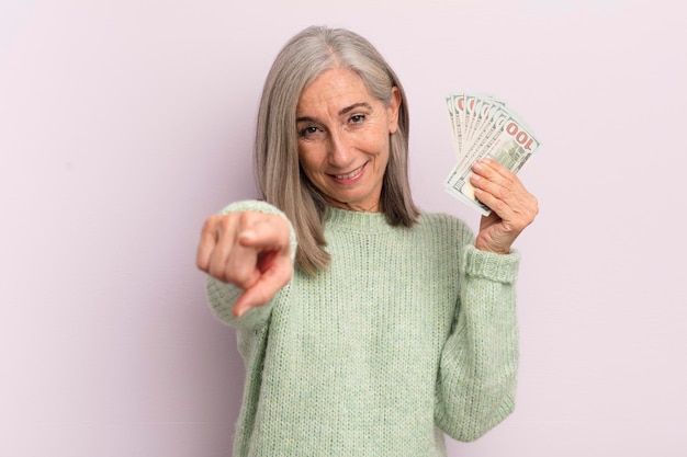 Frau mittleren Alters, die auf die Kamera zeigt und Ihr Dollar-Banknoten-Konzept auswählt