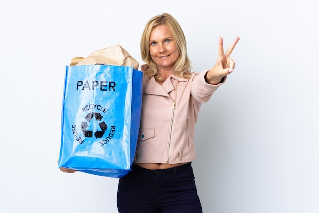 Frau mittleren Alters, die einen Recyclingbeutel voll Papier hält, um lokalisiert auf weißem Lächeln und Siegeszeichen zu recyceln