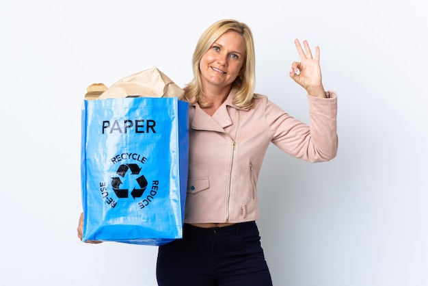 Frau mittleren Alters, die einen Recyclingbeutel voll Papier hält, um lokalisiert auf weißer Wand zu recyceln, die ok Zeichen mit den Fingern zeigt