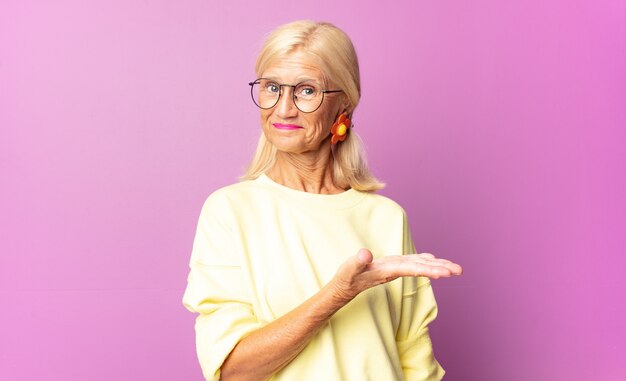 Frau mittleren Alters, die fröhlich lächelt, sich glücklich fühlt und ein Konzept im Kopierraum mit Handfläche zeigt