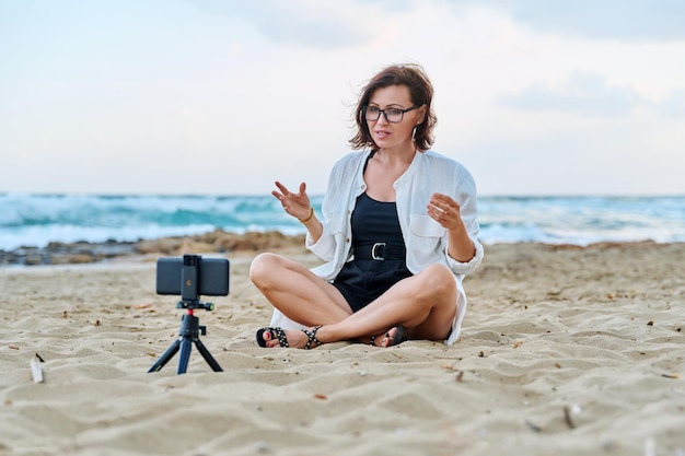 Frau mittleren Alters, die per Videoanruf mit Smartphone am Strand sitzt