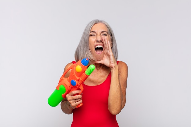 Frau mittleren Alters, die sich glücklich, aufgeregt und positiv fühlt, mit den Händen neben dem Mund einen großen Schrei ausstößt und mit einer Wasserpistole ruft