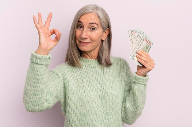 Frau mittleren Alters fühlt sich glücklich und zeigt Zustimmung mit dem Konzept der okay-Geste-Dollar-Banknoten